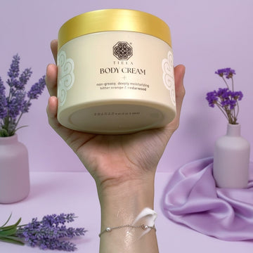 Assemble K - Play .Try .Glow Tiela Perfume Therapy Body Cream Shine 200g in a beige tub with a gold lid, featuring a citrus-floral-cedarwood scent.