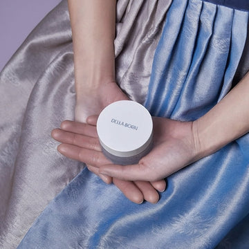 Assemble K - Play .Try .Glow holds a white jar of Della Born Marine Collagen Cream, 50ml, in hands against a blue and silver silk garment.