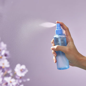 Assemble K - Play .Try .Glow NOT4U Clear Body Mist 200ml, a blue spray bottle held by a hand against a white wall.