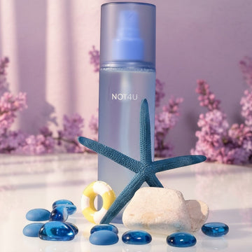 Assemble K - Play .Try .Glow NOT4U Clear Body Mist 200ml, a blue frosted bottle with a spray nozzle, surrounded by blue glass pebbles, a starfish, and a small lifebuoy.
