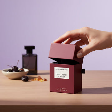 Assemble K - Play .Try .Glow Banana Republic Dark Cherry & Amber perfume box with a hand opening the lid on a table