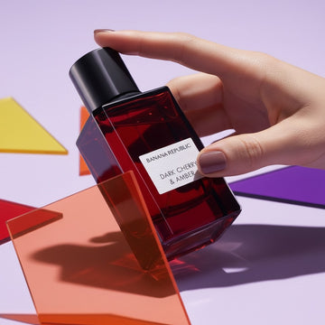Assemble K - Play .Try .Glow Banana Republic Dark Cherry & Amber perfume bottle held by a hand against geometric shapes