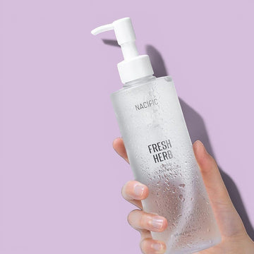 Assemble K - Play .Try .Glow NACIFIC Fresh Herb Origin Cleansing Oil in a clear pump bottle held by a hand.