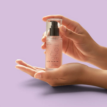 Hands holding Assemble K - Play .Try .Glow Tone Up and Brightening Sunserum in a clear pump bottle against purple.