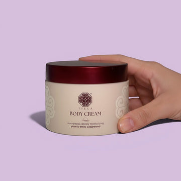 Assemble K - Play .Try .Glow Tiela Body Cream in a cream-colored jar with a dark red lid, featuring a plum and white cedarwood scent.