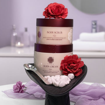 Assemble K - Play .Try .Glow Tiela Perfume Therapy Body Scrub Sunset 250g, a gentle exfoliating sugar scrub with a fruity-floral-woody scent, in a cream-colored jar with a burgundy lid, adorned with red flowers.