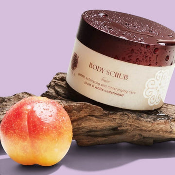 Assemble K - Play .Try .Glow Tiela Perfume Therapy Body Scrub Sunset 250g, a gentle exfoliating sugar scrub with a fruity-floral-woody scent, in a cream-colored jar with a burgundy lid, adorned with red flowers.
