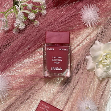 Assemble K - Play .Try .Glow Inga Water Double Lasting Scented Tint in Tuberose shade, a rosy pink lip tint with a clear glass bottle and pink cap, surrounded by pink decorative grass and flowers.