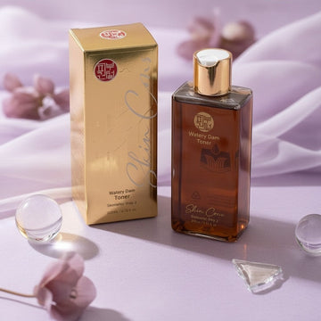 Assemble K - Play .Try .Glow Skinterior Step 2 Watery Dam Toner in a brown glass bottle with gold accents, on a light surface with water ripples.